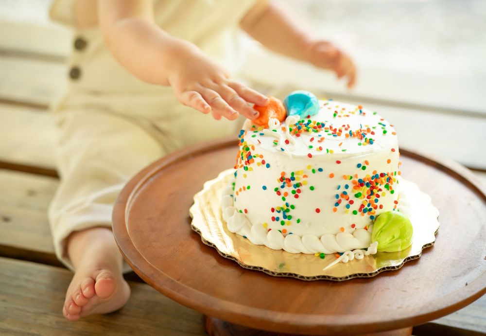 Small hands reach for a white birthday cake decorated with colorful sprinkles on a wooden table.