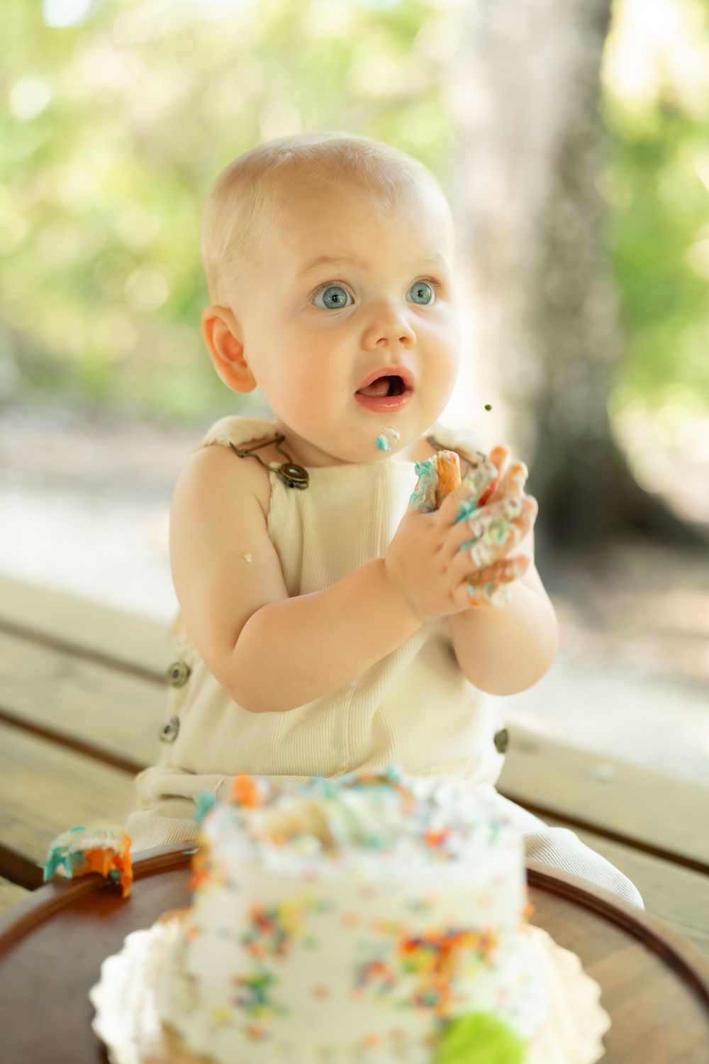 A baby in cream overalls enjoying cake with white frosting and sprinkles outdoors.