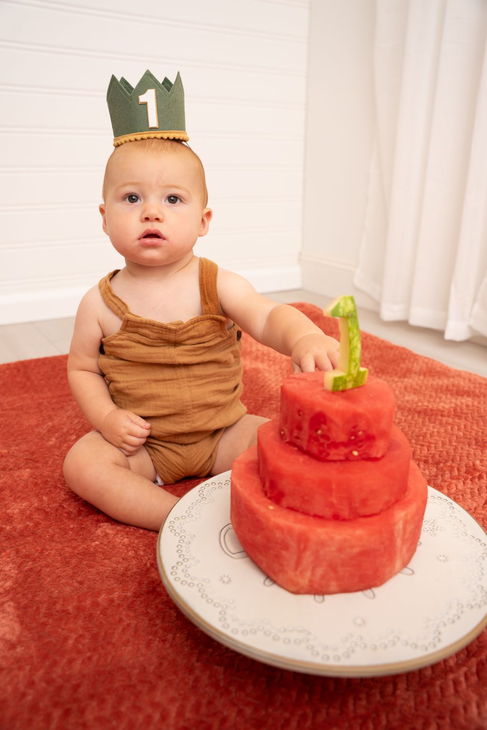 A baby wearing a crown and brown romper sits beside a watermelon cake on a red rug.