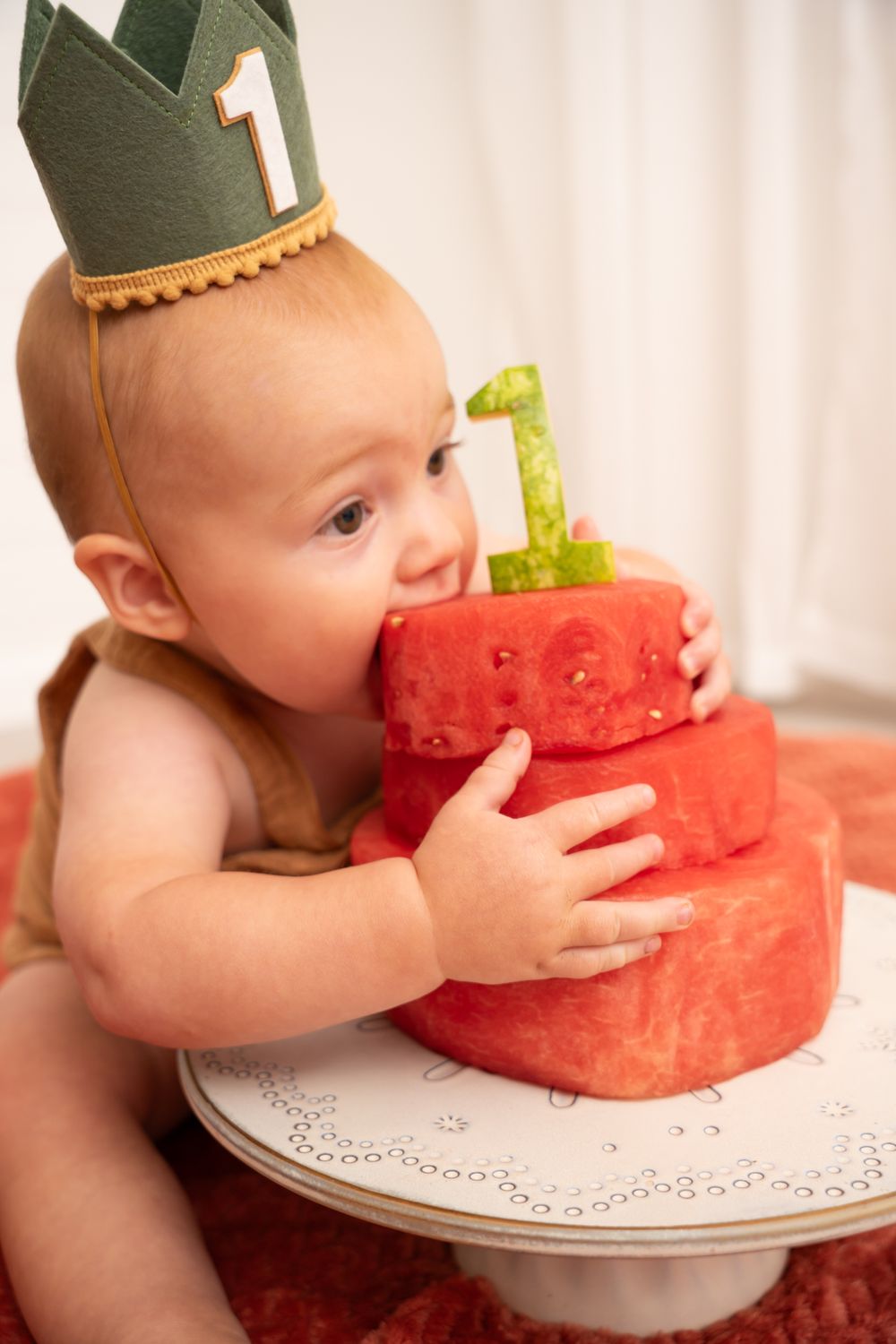 A baby wearing a green crown eagerly bites into a tiered watermelon cake.