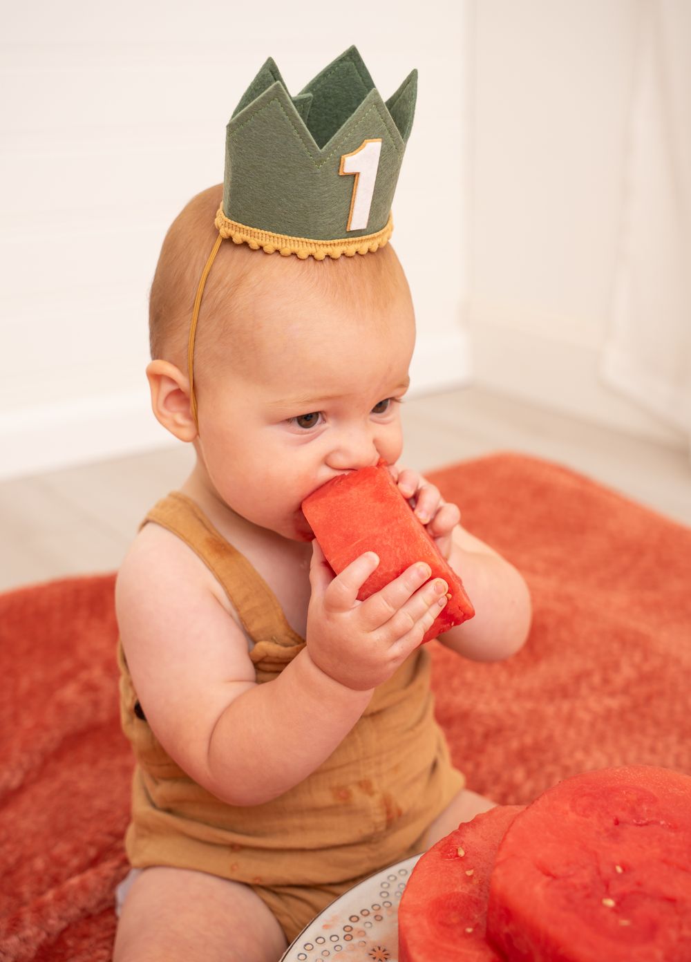 A small child in a brown outfit and green crown nibbles on a piece of watermelon cake.