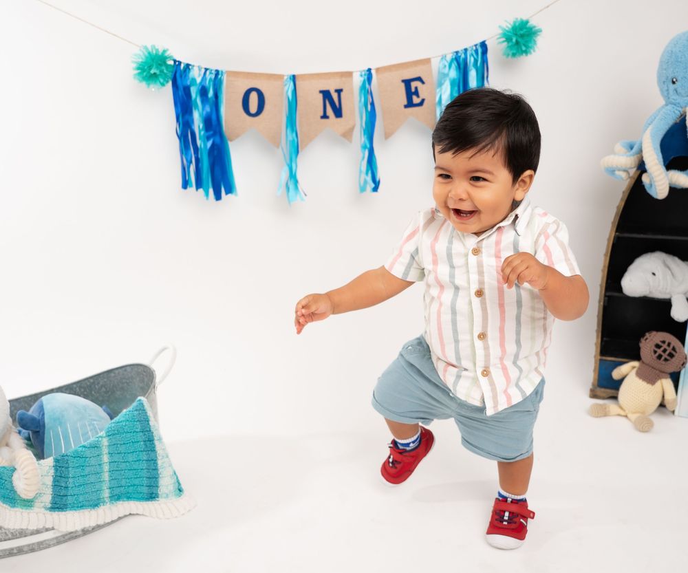 A toddler in a striped shirt and blue pants dances joyfully under a blue 'ONE' banner with teal pom poms.