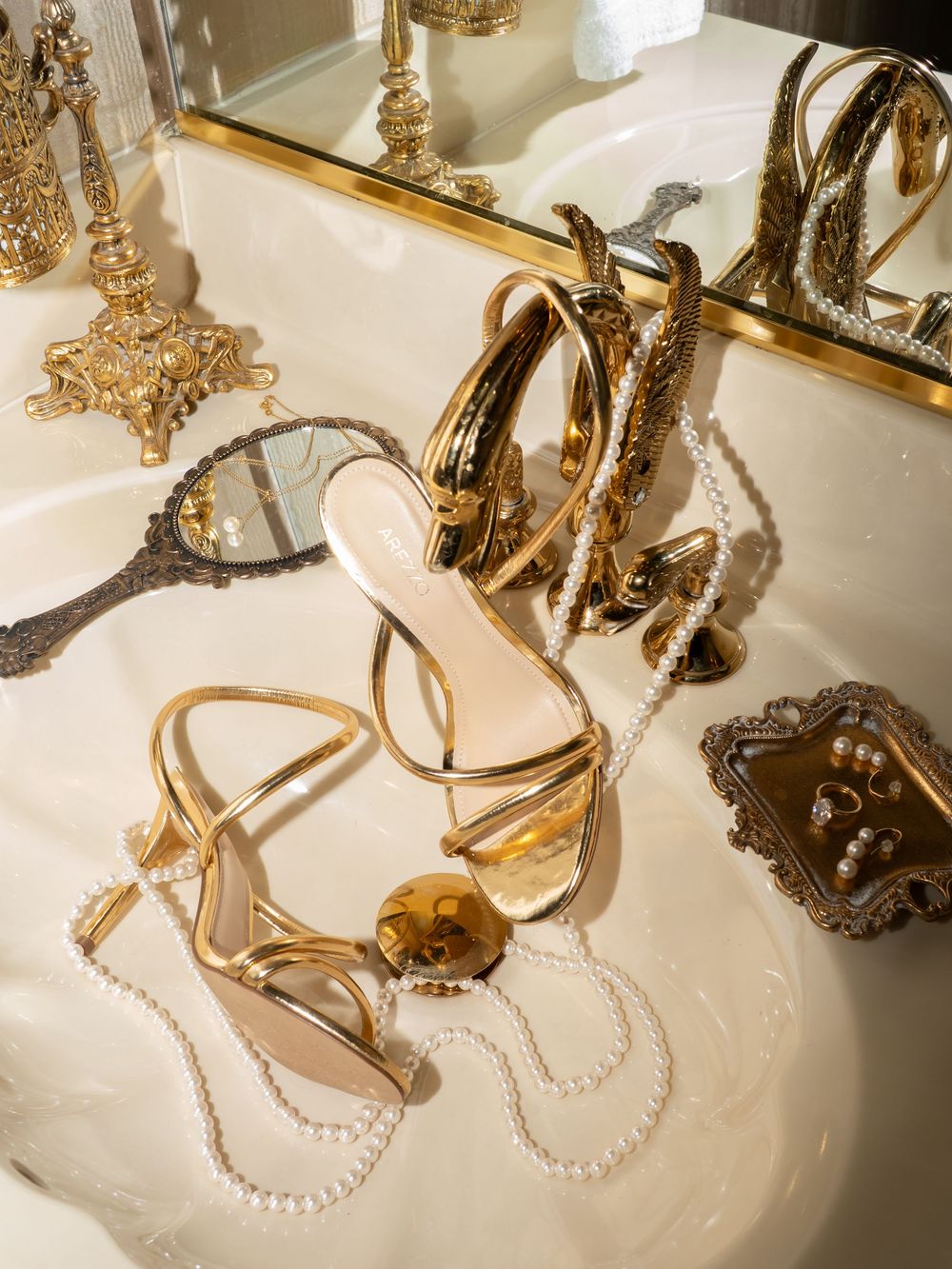 Elegant gold strappy sandals and pearl necklaces displayed on a white surface with reflective mirror accents.