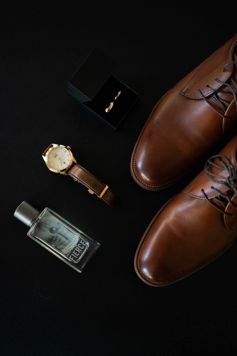 Brown leather dress shoes with watch and cologne on black background.