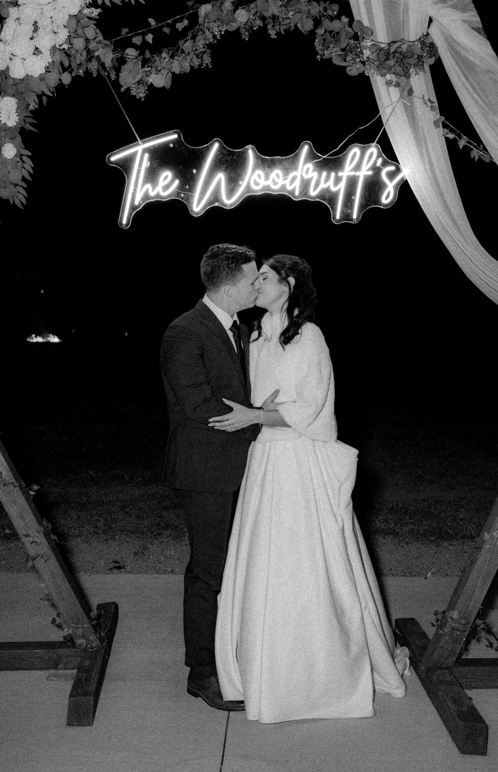 Wedding couple under illuminated neon sign reading 'The Woodruffs' with romantic lighting.