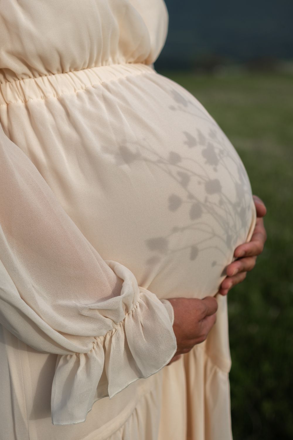 Close-up of pregnant belly in flowing white maternity dress against natural backdrop.