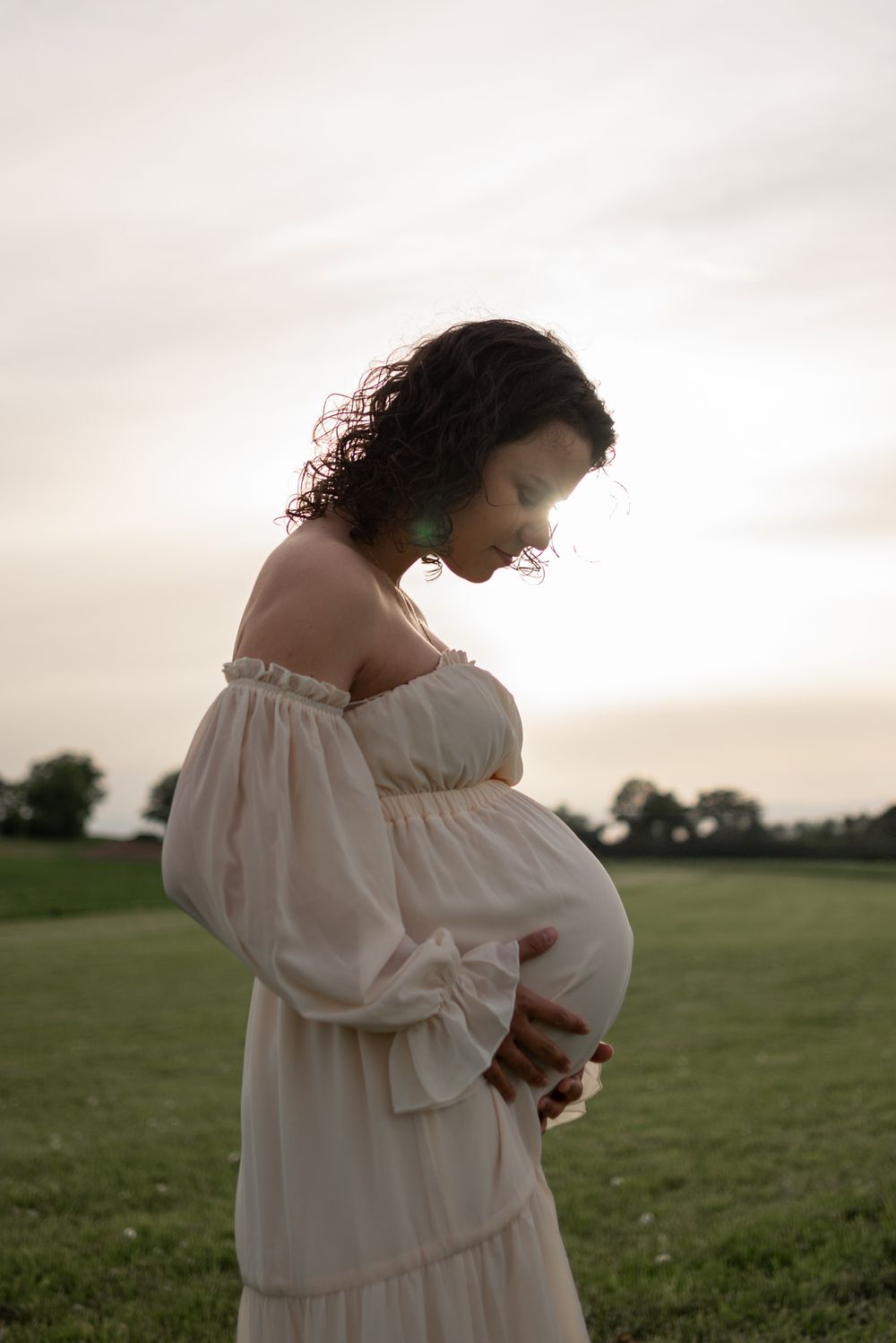 Expectant mother in flowing white dress standing in field at sunset.