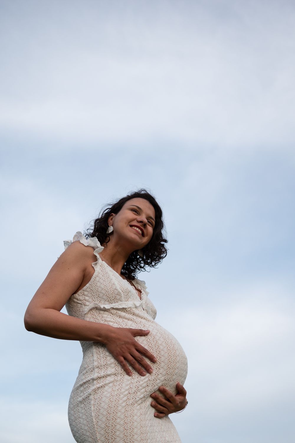 Joyful maternity moment captured against blue sky in white lace dress.