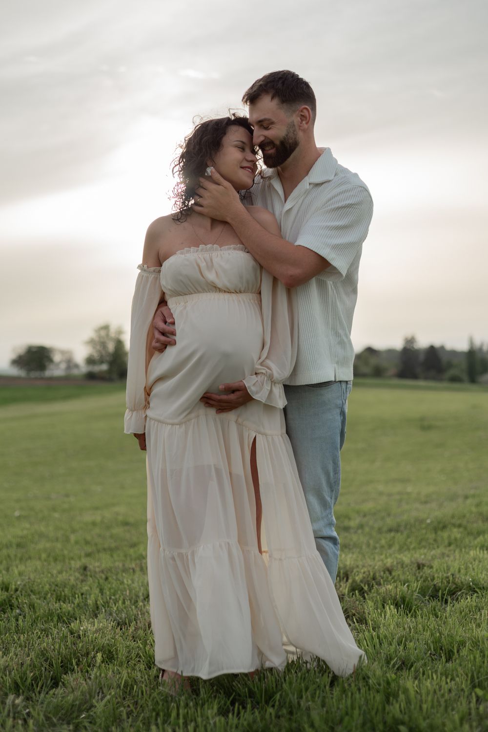 A romantic couple embraces in a flowing white dress against a green meadow at sunset.