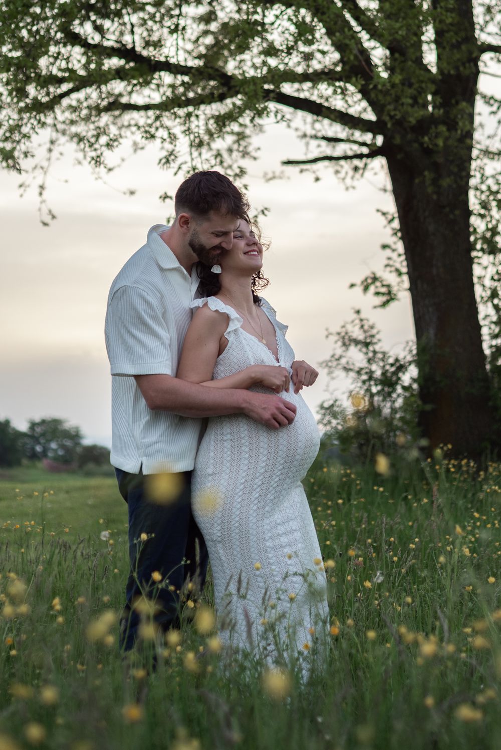 A couple shares an intimate moment in a wildflower meadow under large trees.