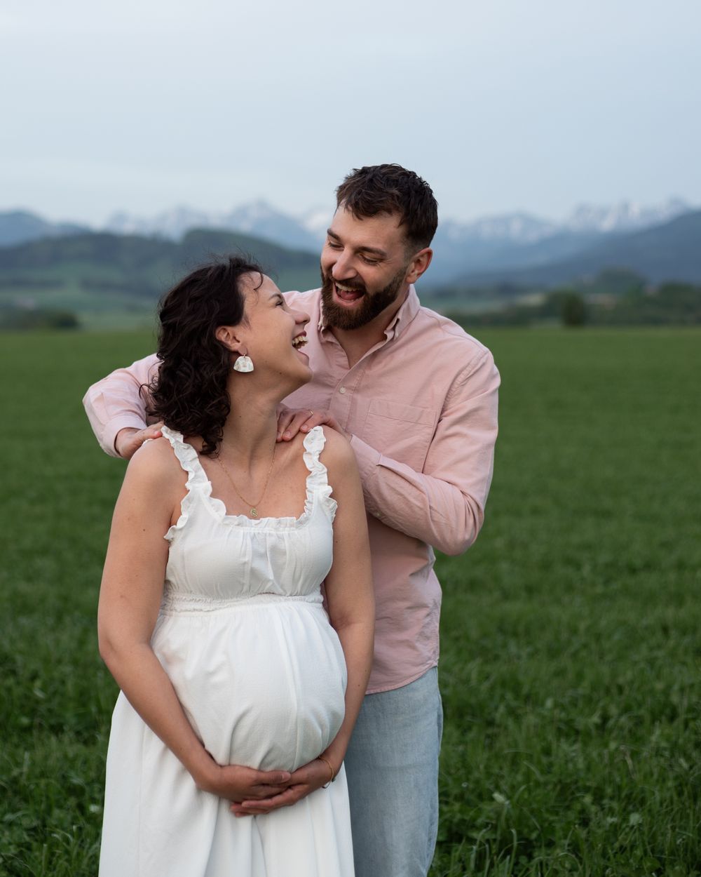 A couple in white and pink clothing embraces in front of mountain scenery.