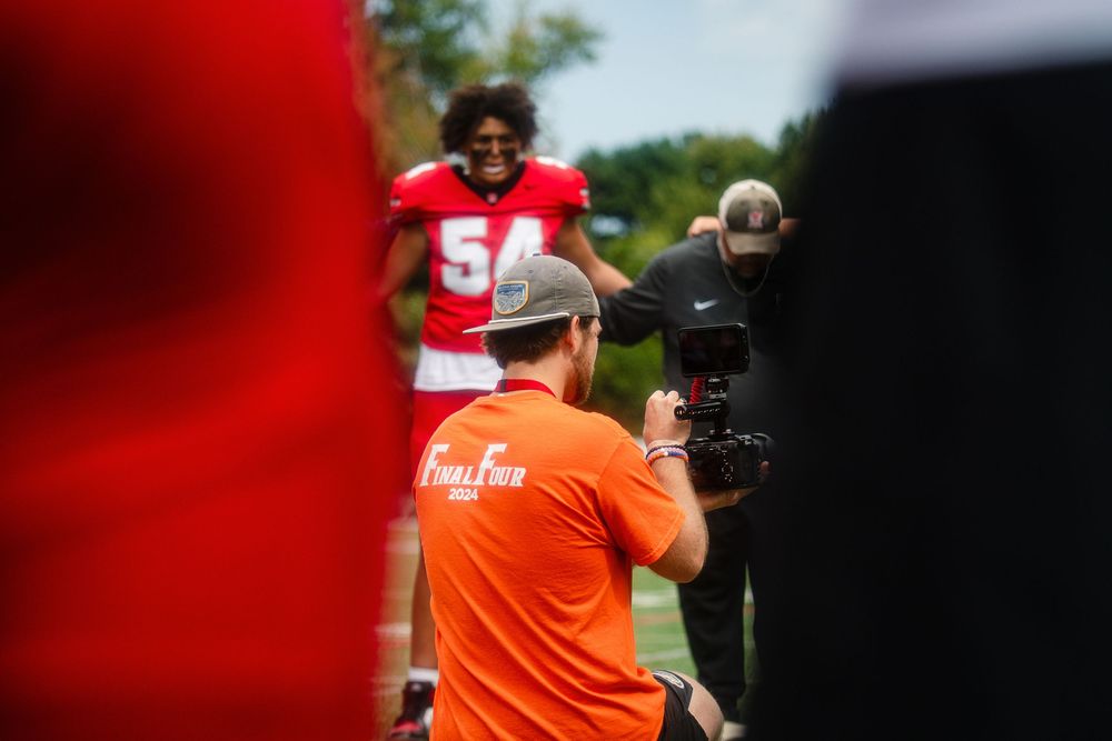 Football player in red jersey number 56 being filmed by camera operator in orange shirt during outdoor practice session.
