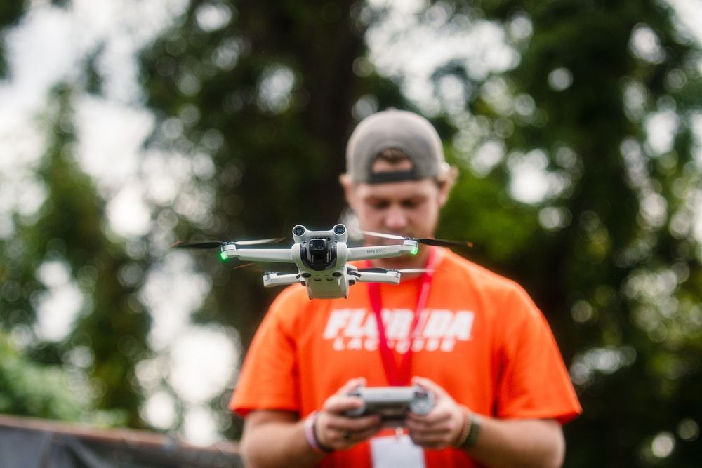 A person in an orange shirt operates a drone remote control while standing outdoors.