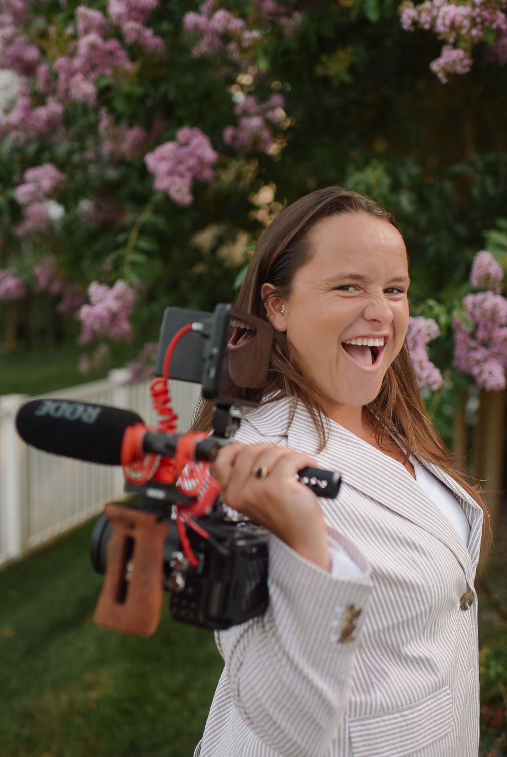 Someone holding a professional video camera laughs joyfully near blooming pink flowers in a garden.