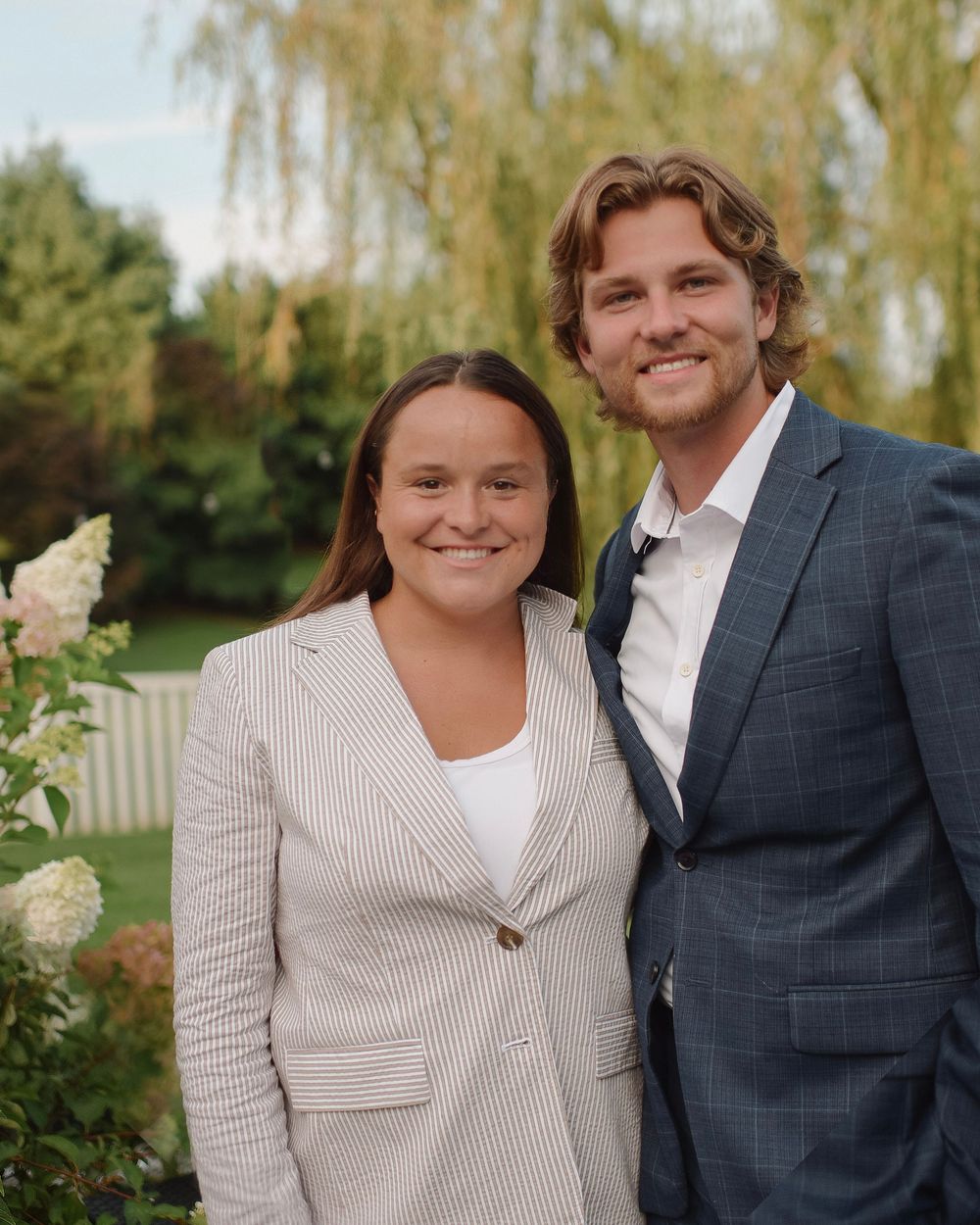 Happy couple posing for outdoor engagement photos by willow trees in a garden setting.