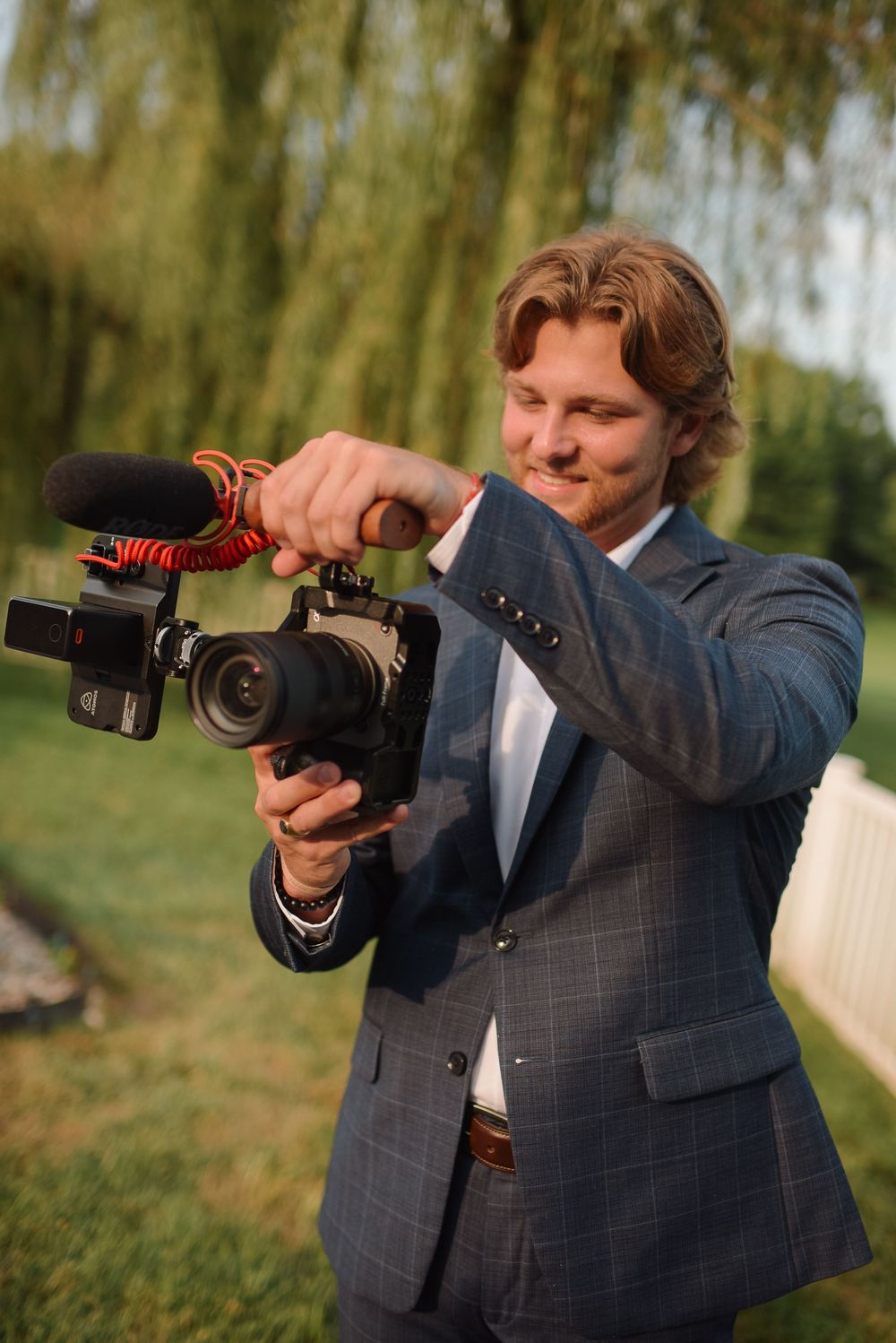 Professional videographer wearing gray suit adjusts settings on professional video camera outdoors near willow trees.