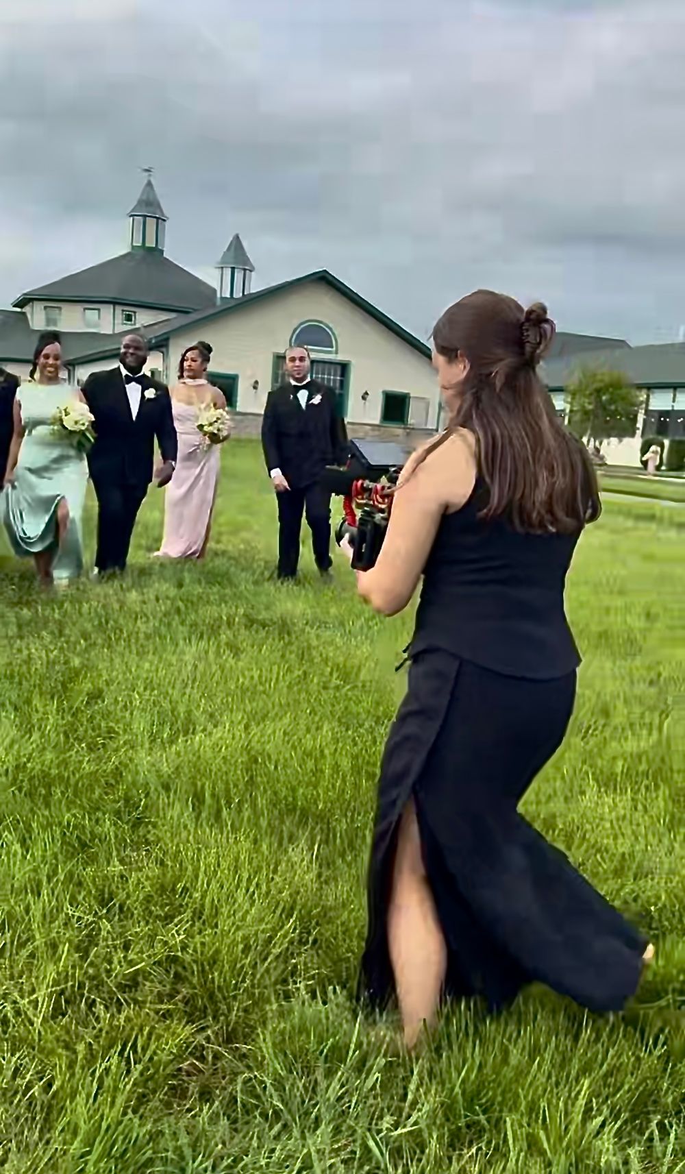 A photographer in a navy dress takes pictures of wedding party members on grass with a church in the background.