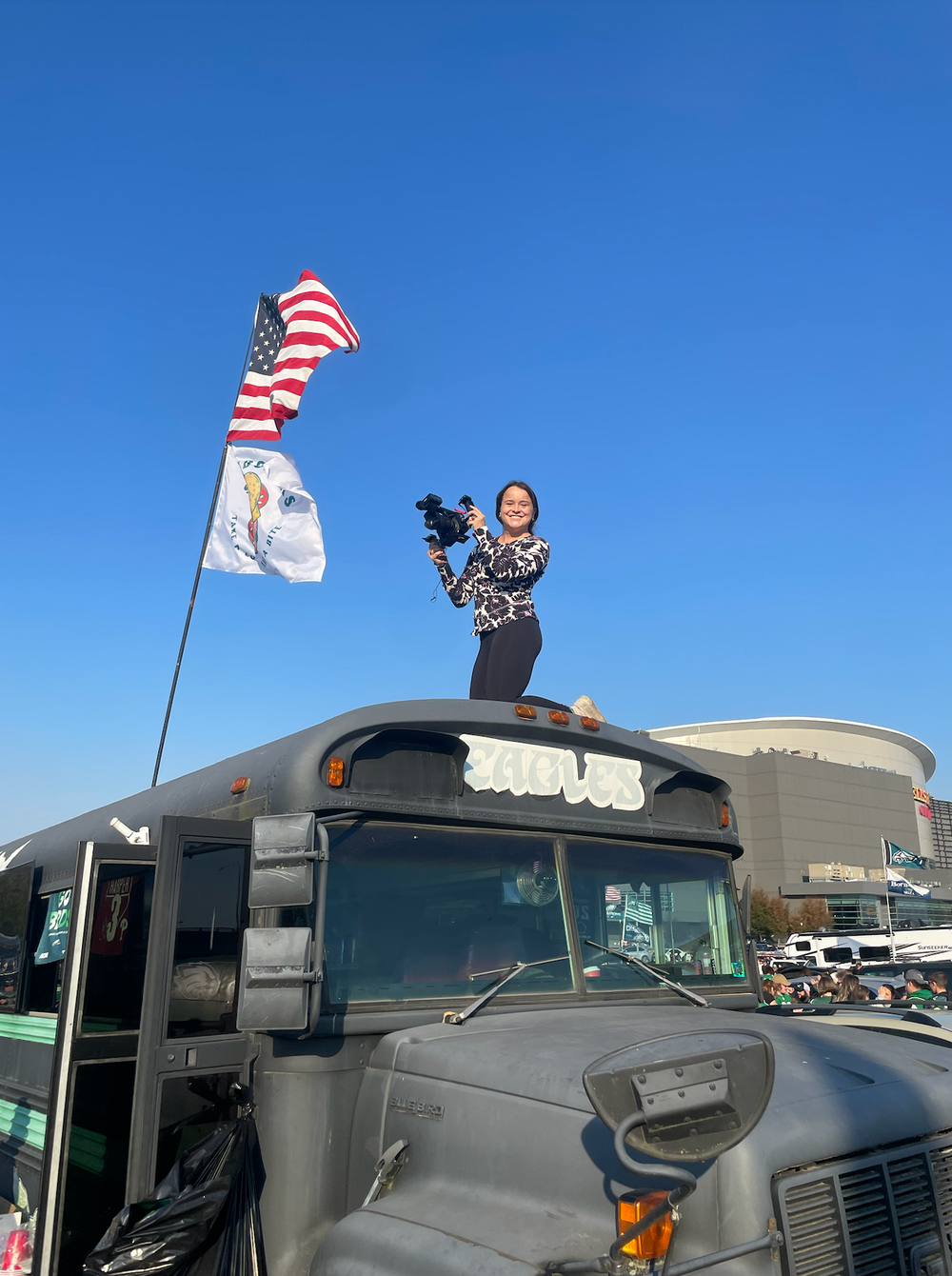Person filming with camera on top of school bus with American flag waving.