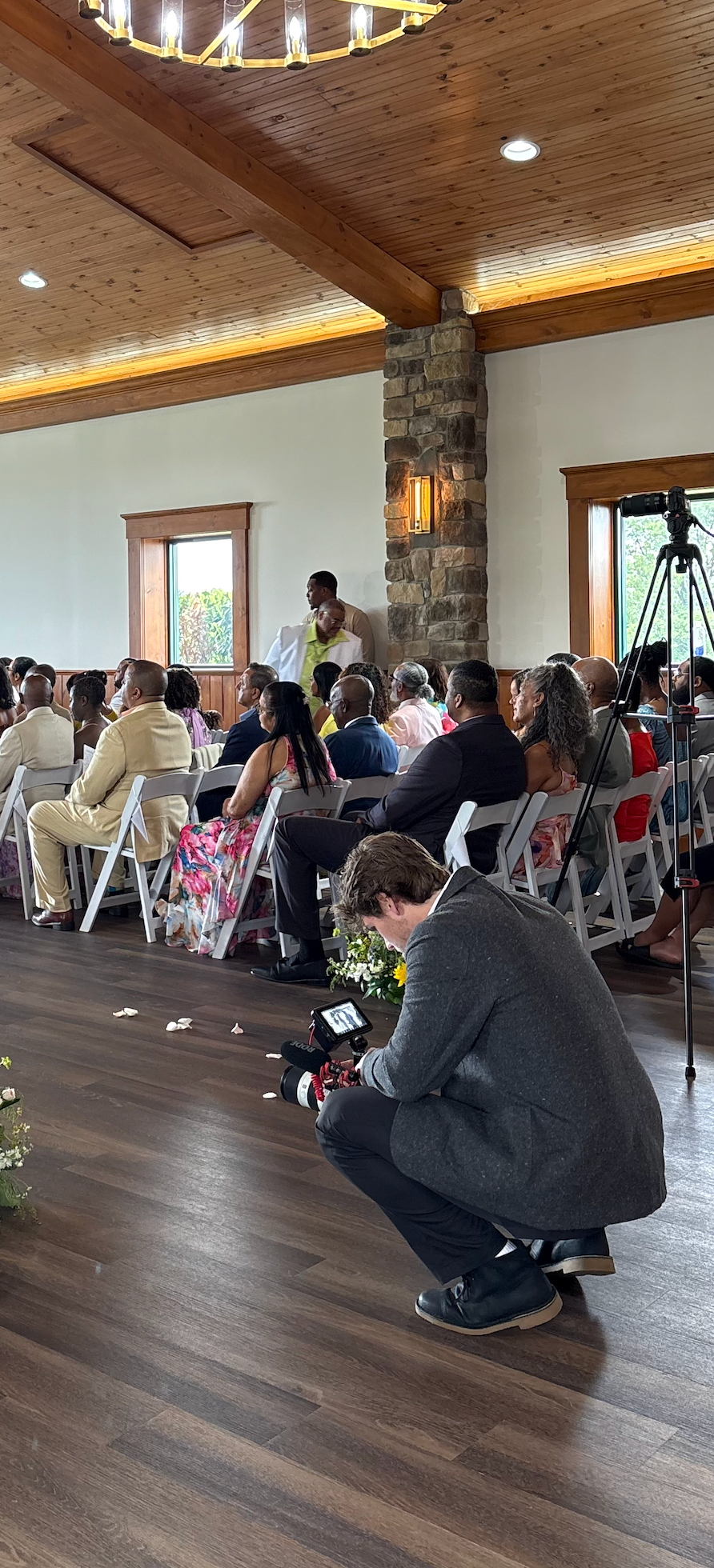 Event photographer crouches on hardwood floor while attendees sit in folding chairs in a rustic venue with wooden beams.