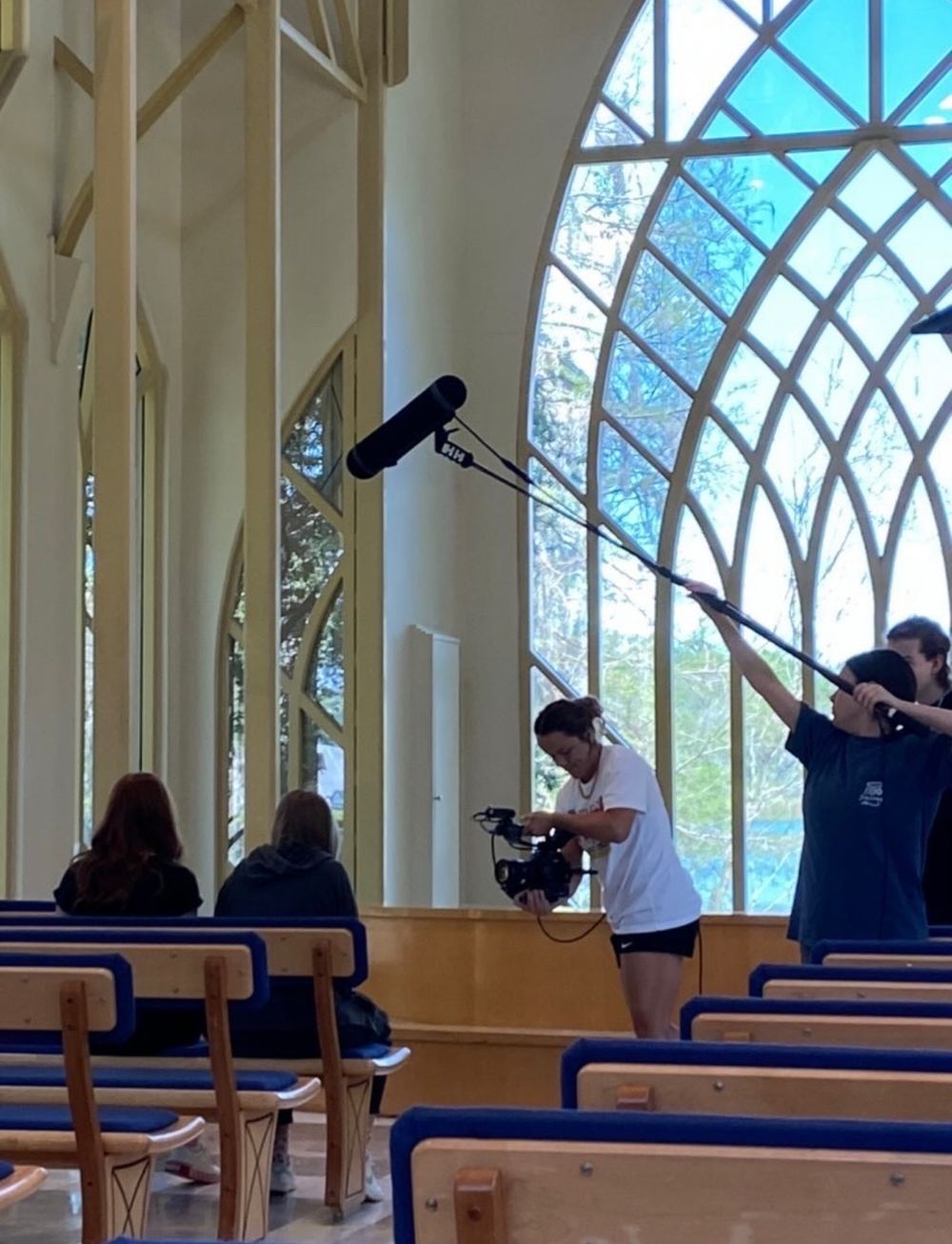 Film crew recording with boom mic in church or chapel with arched windows.