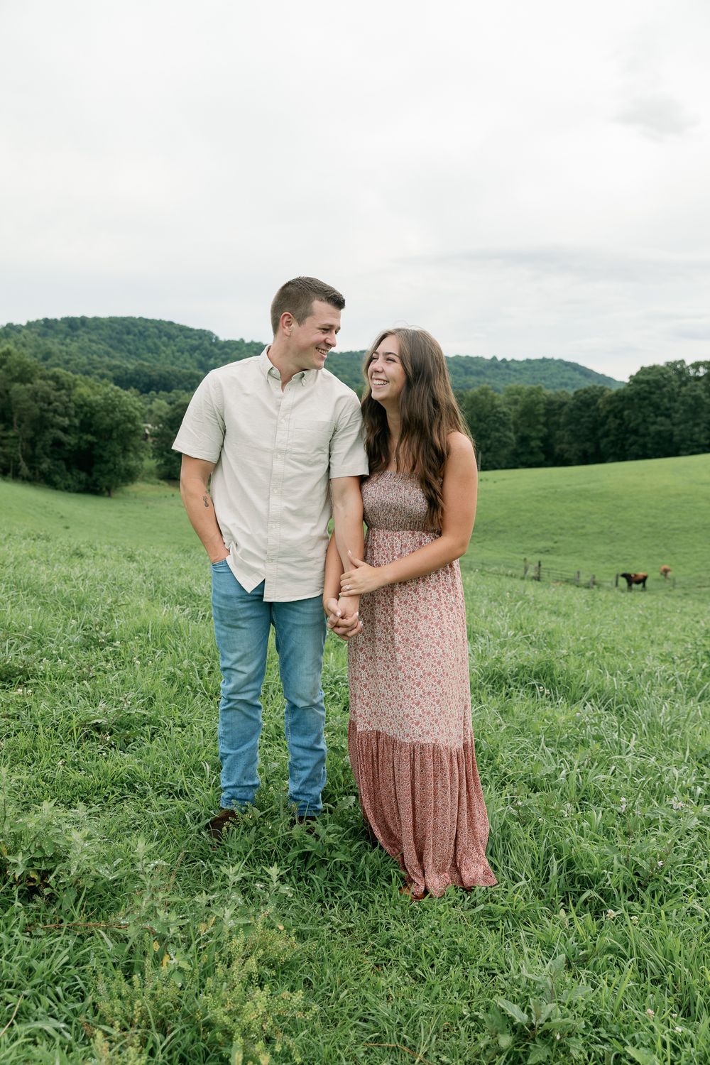 A couple stands together in a green summer meadow with rolling hills in the background.