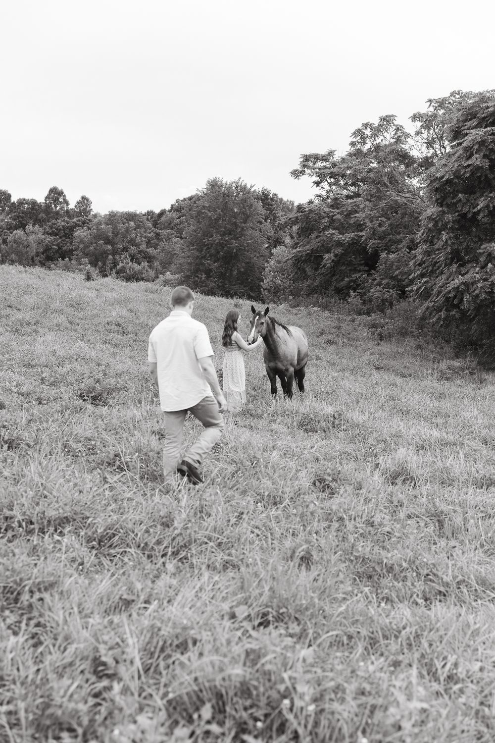 Black and white photo of a couple walking through a grassy field with horses in the distance.
