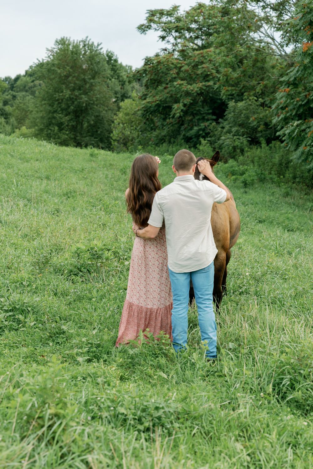 A couple walks through tall grass towards horses grazing in a pastoral field.