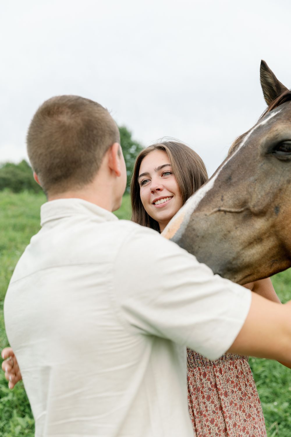 A couple interacts with a curious brown horse in a sunny meadow.