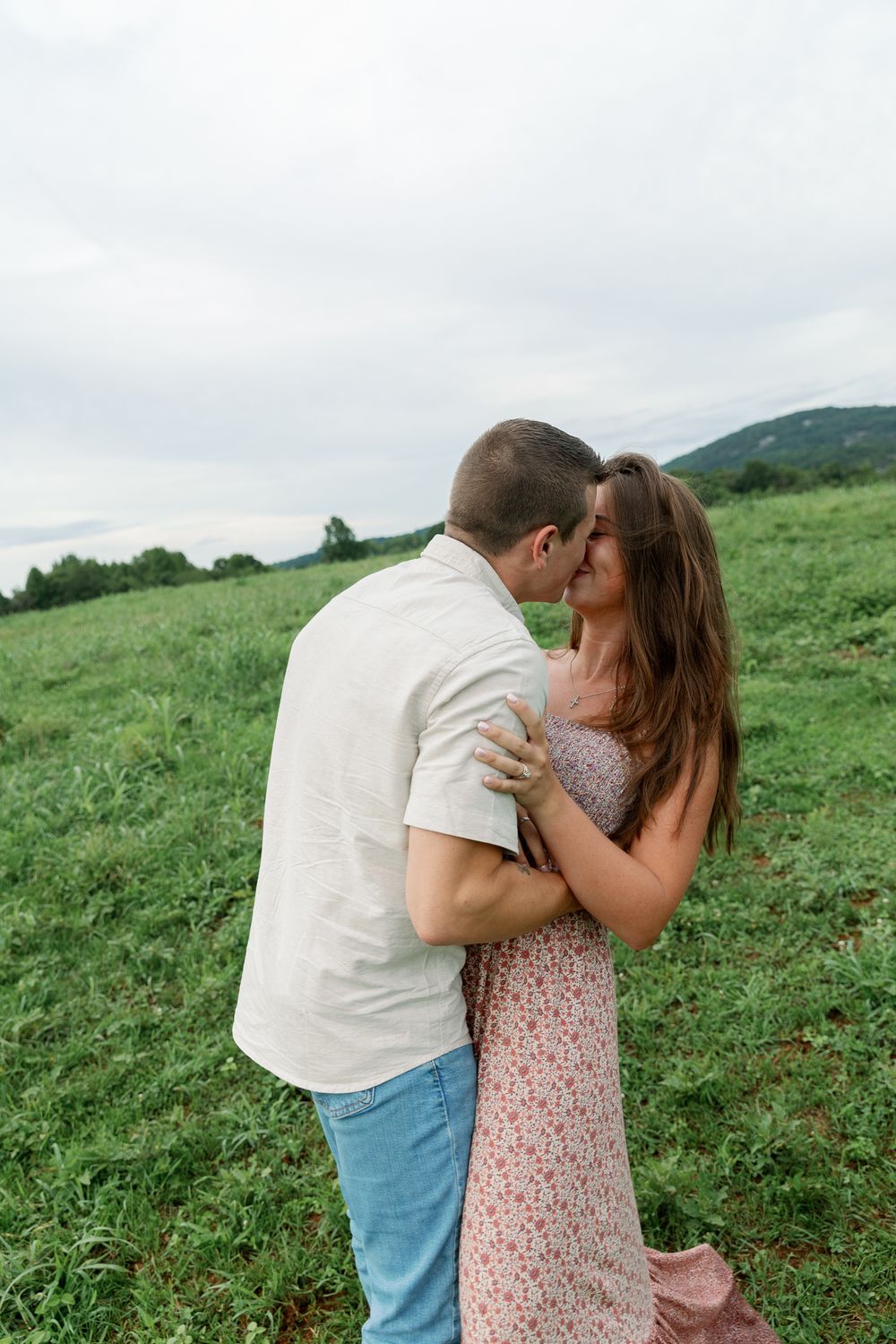 A couple embraces in a scenic hilltop meadow on a cloudy summer day.