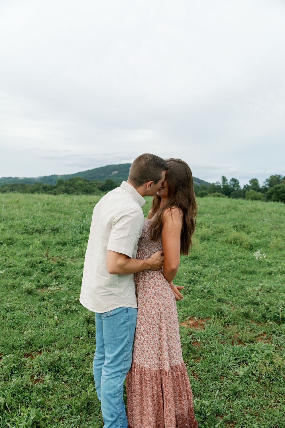 A couple shares a kiss while standing in a verdant summer meadow.