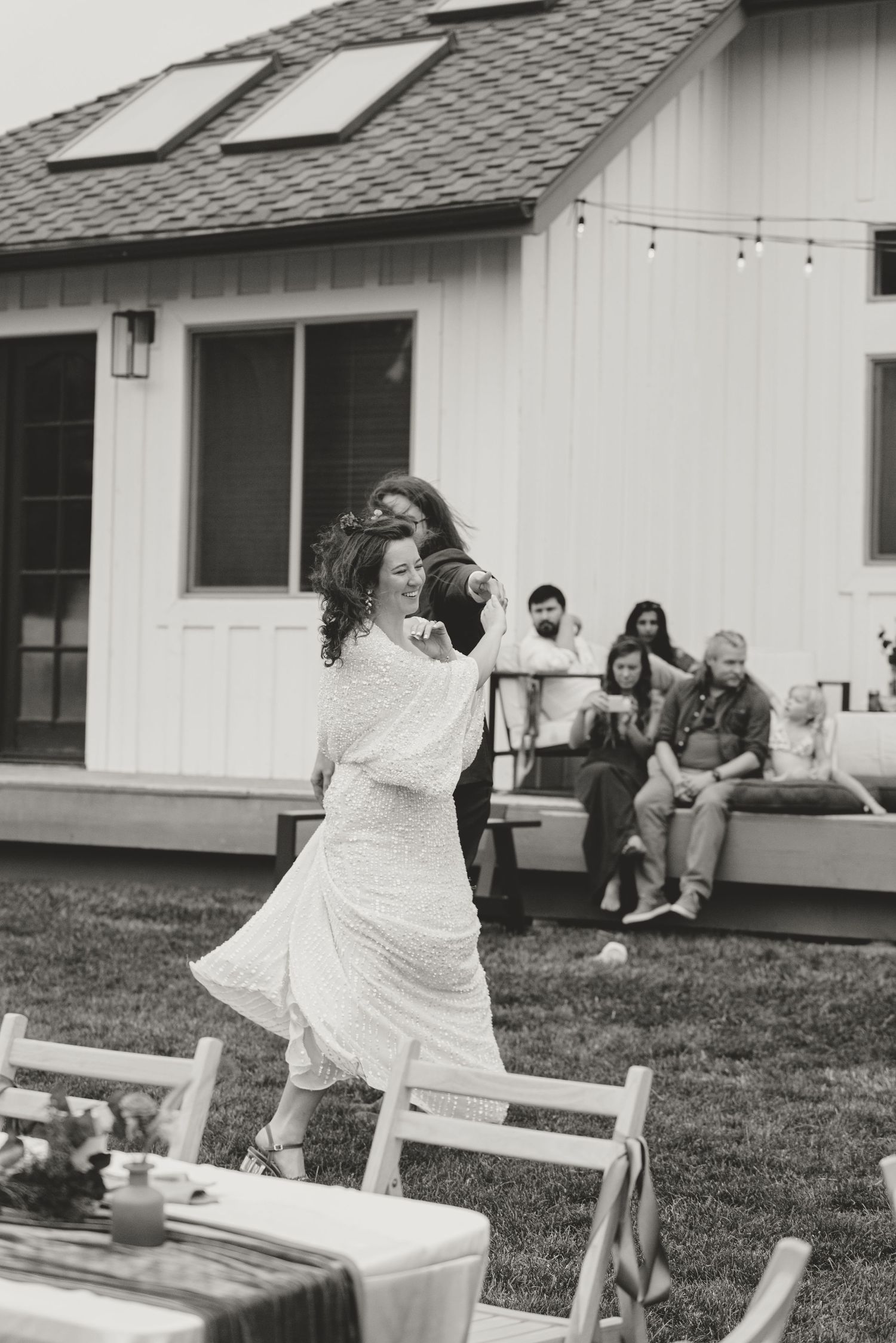 Person in white dress dancing at outdoor gathering with house and people in background.