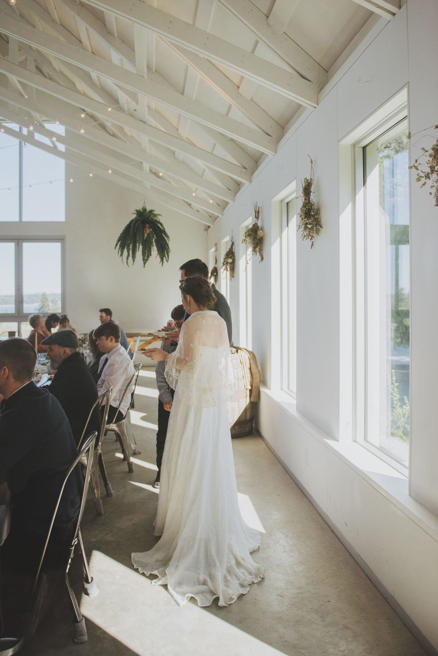 Wedding guests seated at tables while bride stands near bright windows in an elegant white venue.