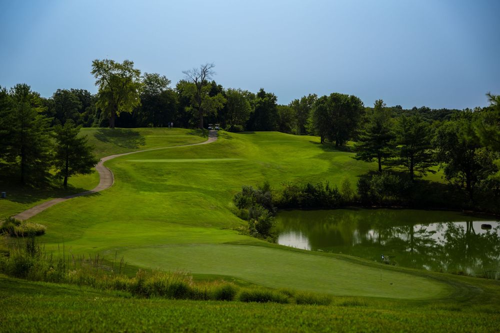 A scenic golf course hole with rolling green fairways and a small pond surrounded by trees.