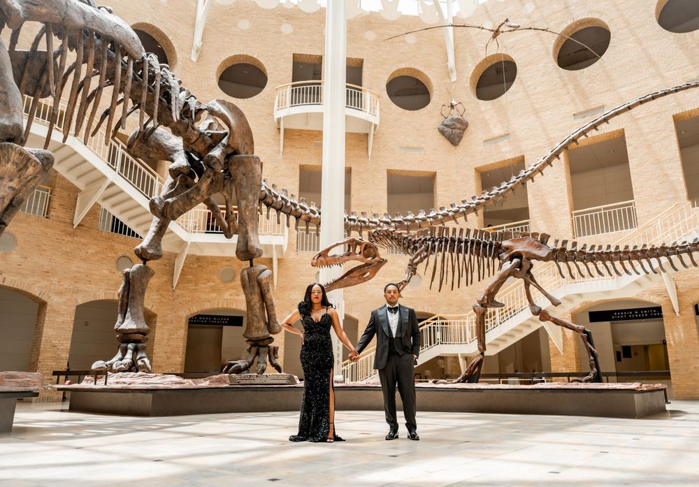Couple poses dramatically among dinosaur skeleton displays in natural history museum.
