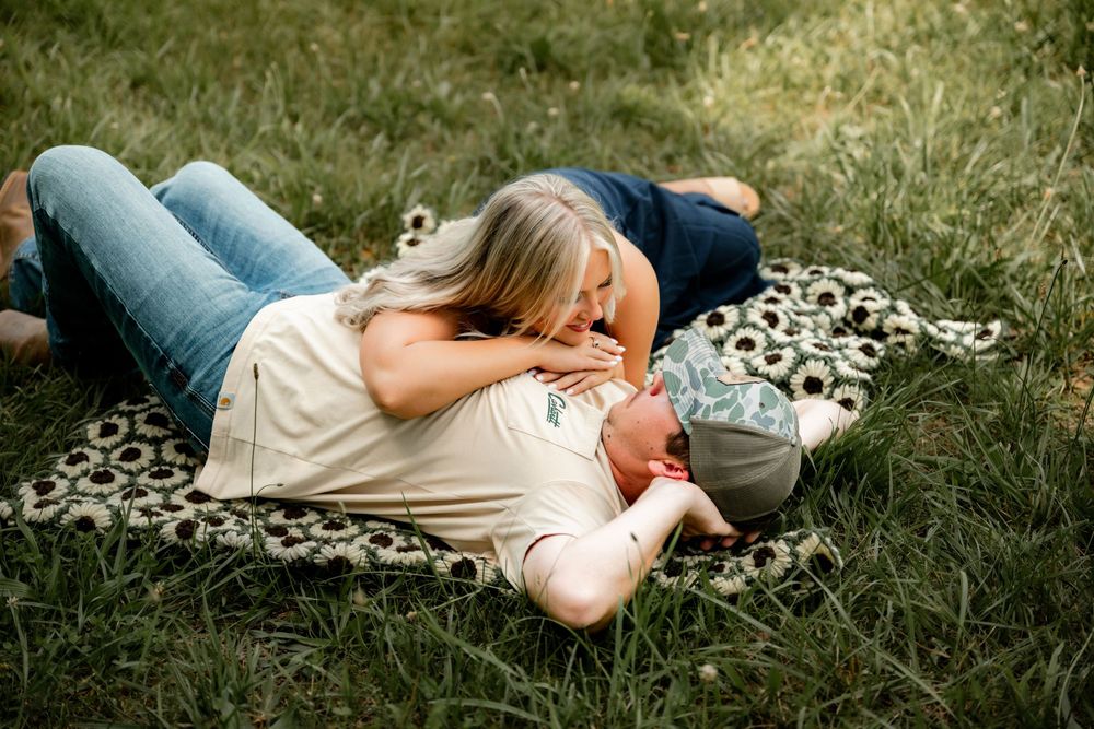 Couple laying on a floral blanket in green grass, woman leaning over man in a tender romantic outdoor moment.