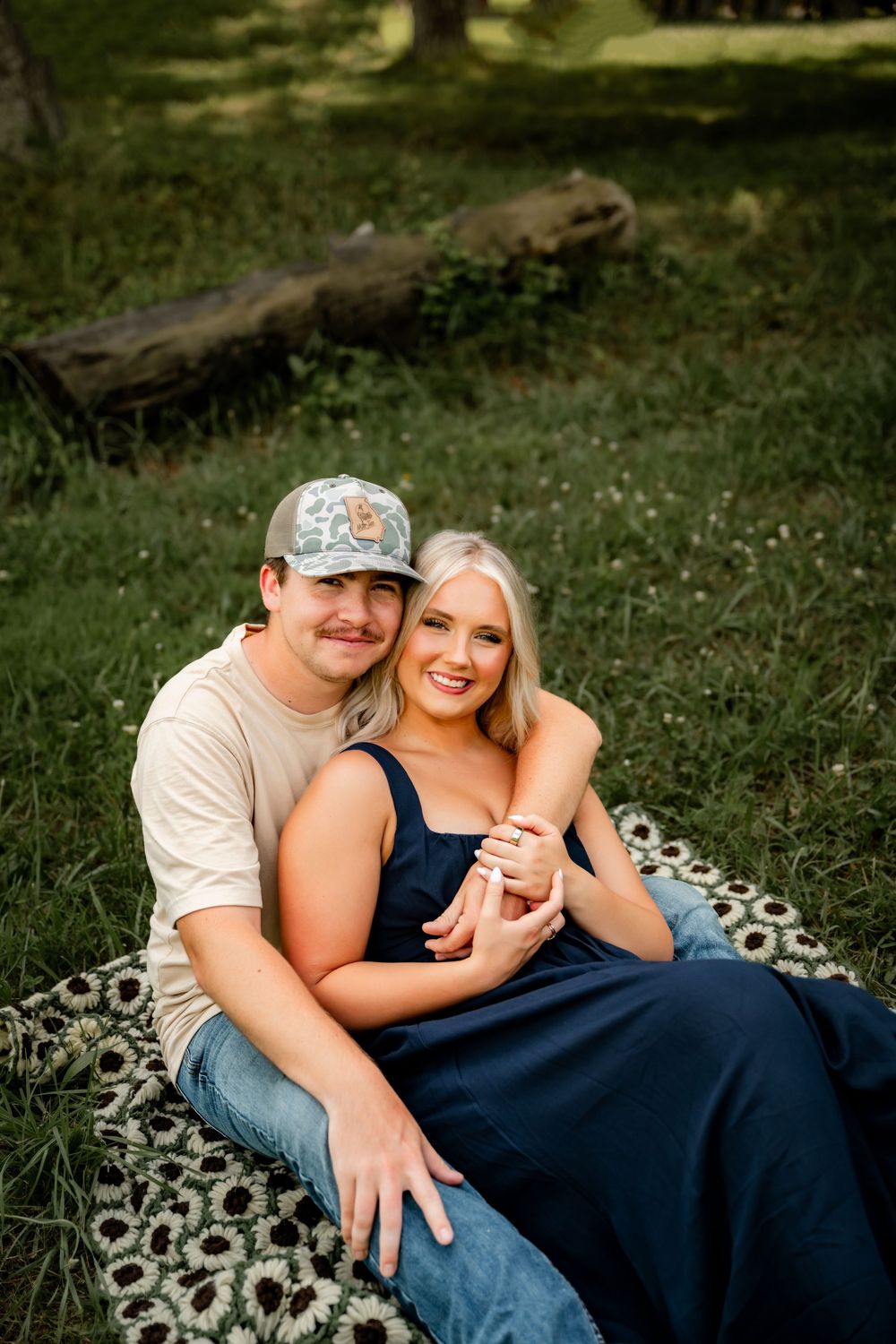 A couple sits together on a floral blanket in a lush green park, smiling warmly at the camera.