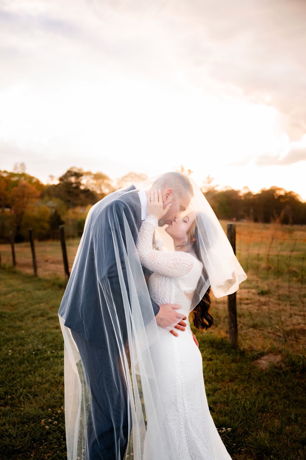 Bride and groom sharing a romantic kiss under a flowing veil at golden hour in a rustic outdoor setting.
