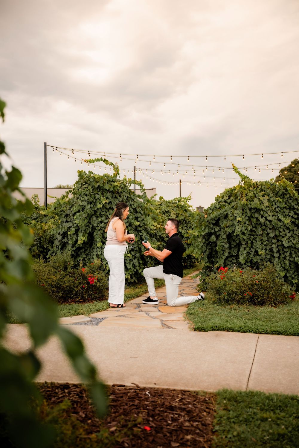 Man kneeling on one knee proposing to woman in white outfit in lush garden with string lights overhead.