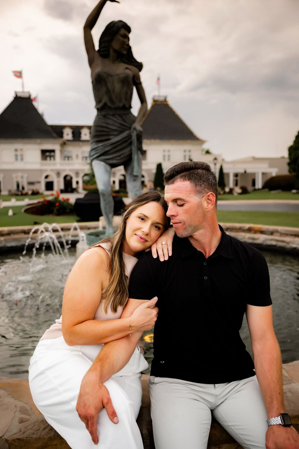Couple sitting by a fountain with a bronze dancer statue and elegant white mansion in the background.