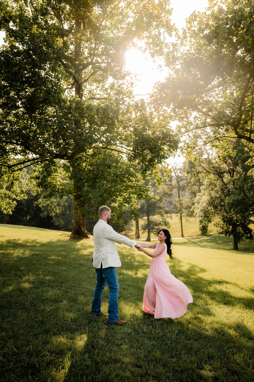Couple dancing in golden sunlight on lush green grass, woman in flowing pink dress, man in white jacket and jeans.