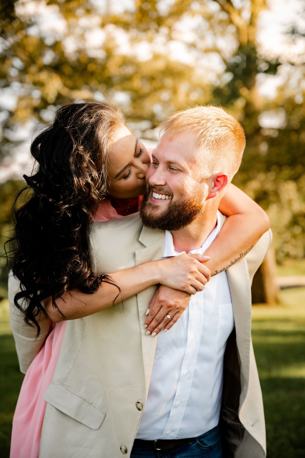 A joyful couple embraces outdoors, woman kissing mans cheek as he smiles broadly, set against lush green trees.