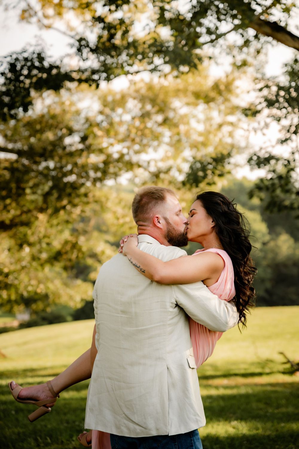 Couple sharing a romantic kiss in a lush green park, man lifting woman in pink dress, surrounded by golden sunlit trees.