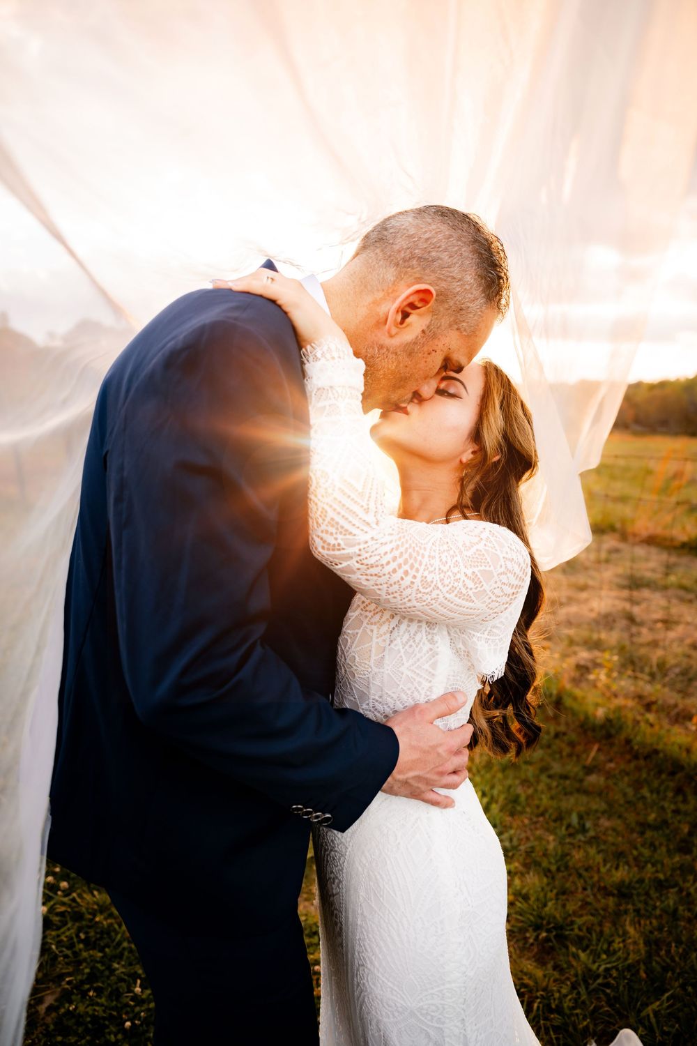 A bride and groom share a romantic kiss under a flowing veil at golden hour in an outdoor field.