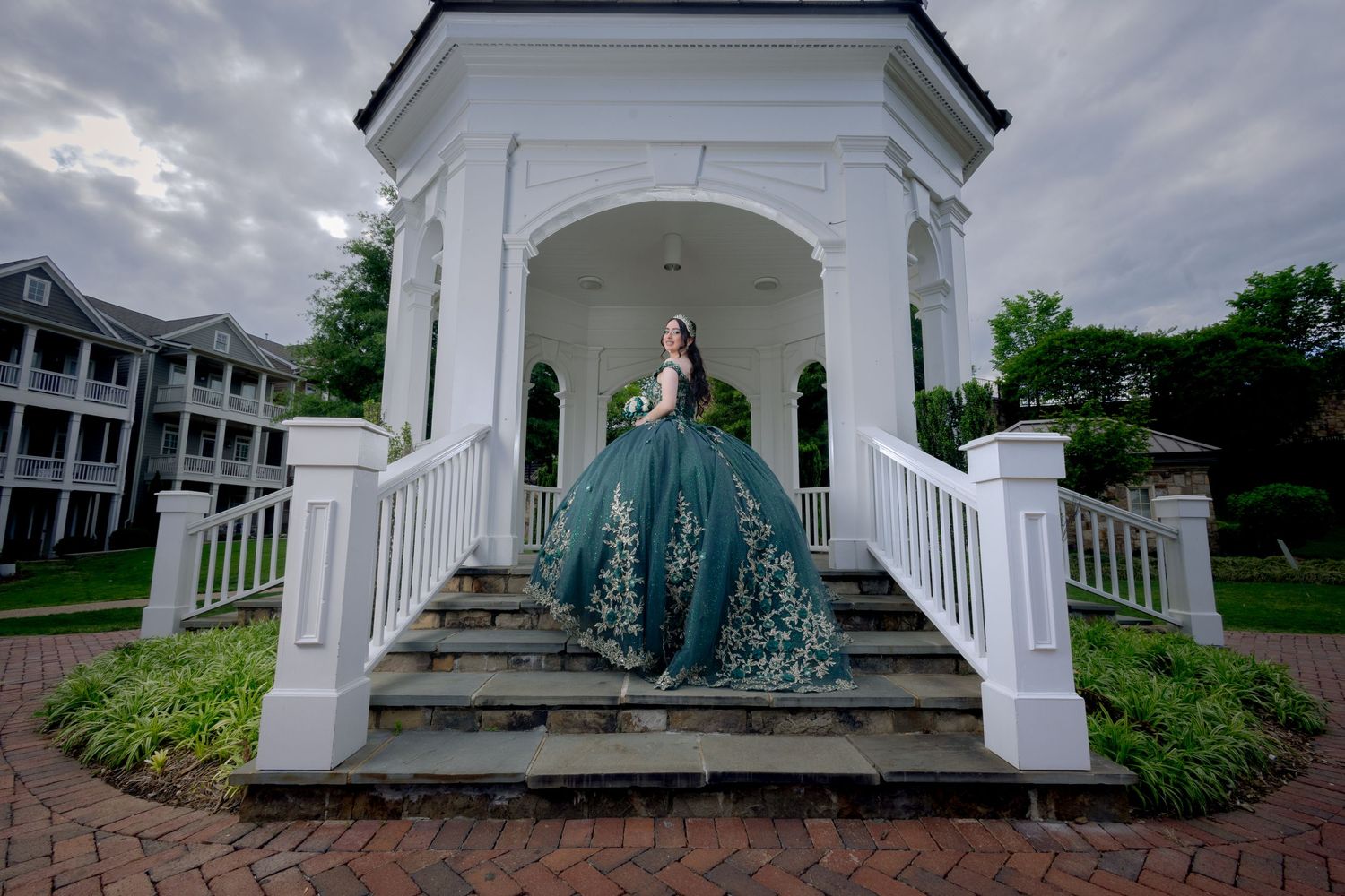 Elegant teal ball gown displayed in a white classical gazebo with colonial architecture.