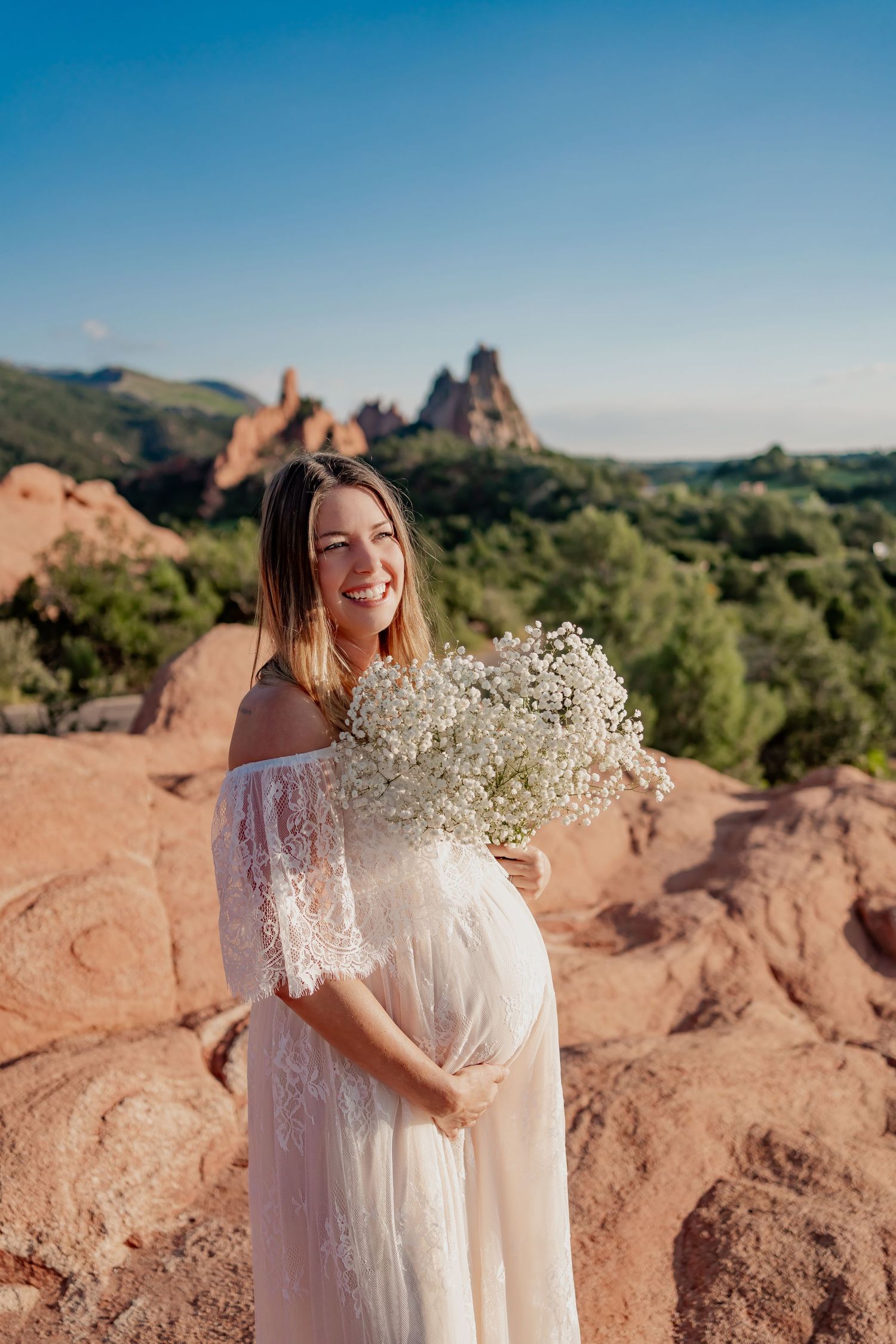 Outdoor maternity photo in white lace dress holding baby's breath bouquet against red rocks.