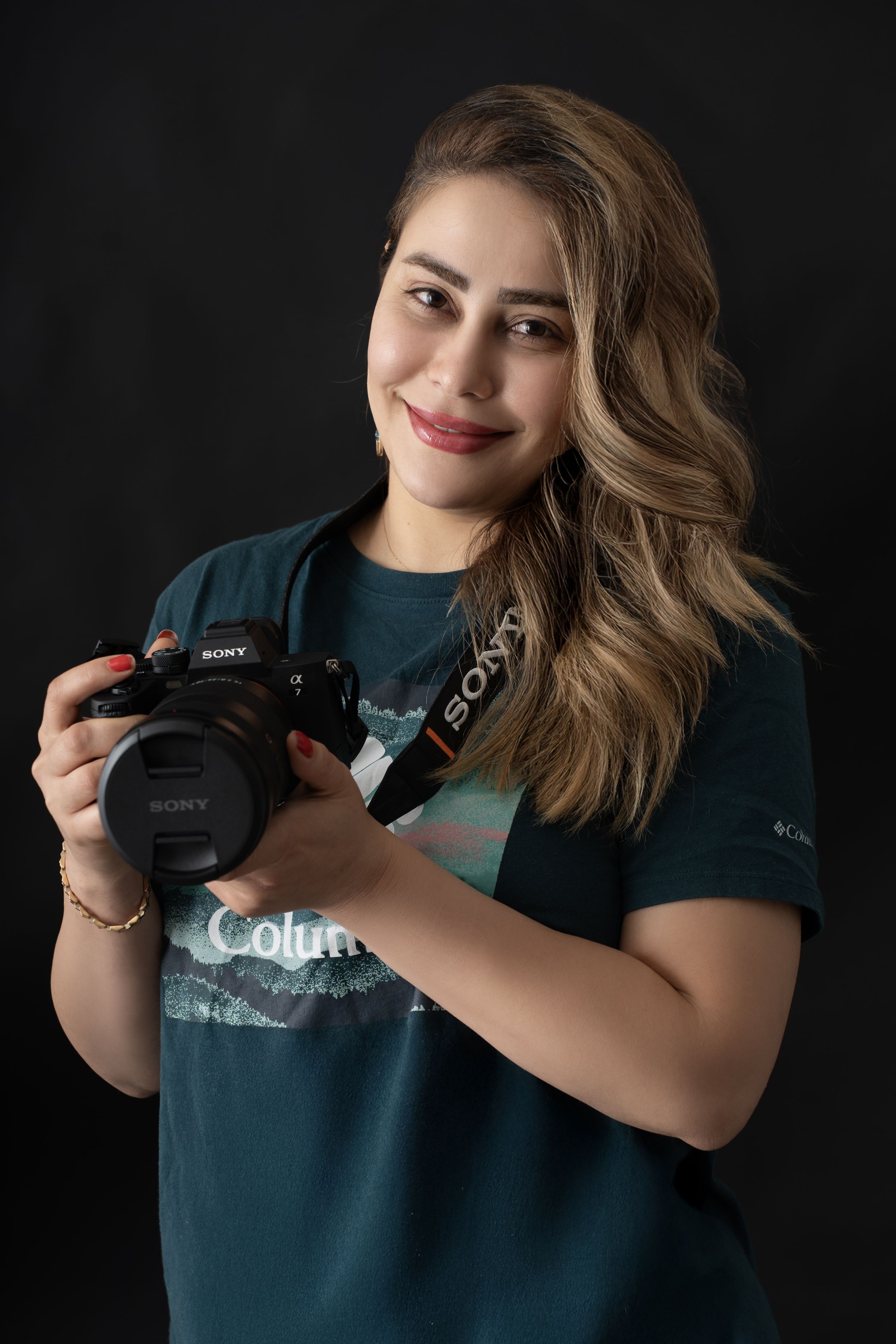 Young woman smiling and holding a Sony camera, wearing a teal Columbia t-shirt against a dark background.