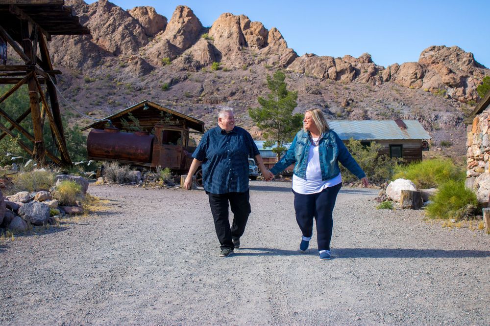 Couple walking hand in hand on a gravel path with rocky mountains in background.