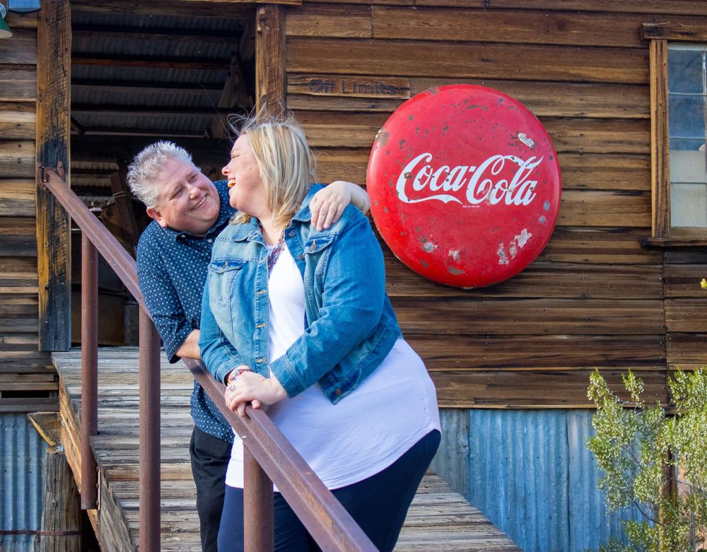 Couple laughing together on rustic wooden steps beside a vintage Coca-Cola bottle cap sign.