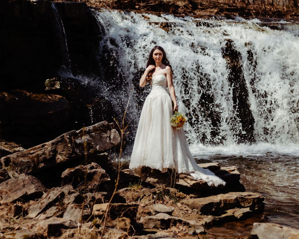 A bride stands gracefully by a cascading waterfall holding a small bouquet.