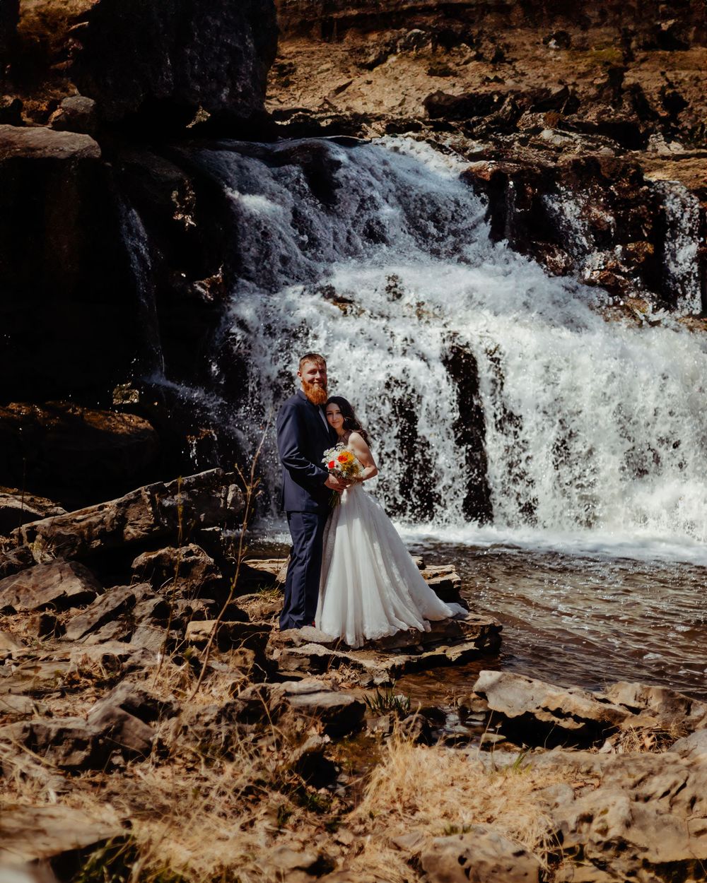 Wedding couple embraces near a flowing waterfall on rugged rocks during their photoshoot.